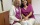 Frau Mauksch und Frau Röth sitzen nahe bei einander und lächeln in die Kamera. Beide tragen die lila-farbene Praxisuniform mit weißen Hosen. Im Hintergrund ein geschlossener Aktenschrank mit Pokalen oben auf und Photographien an der Wand